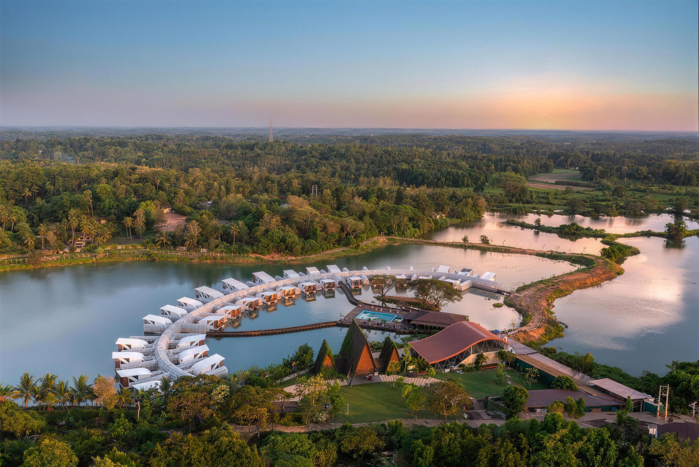 Aerial view of Bolagala at dusk: white floating cabanas in an arc on the lake, with the pool and pavilions on the shore